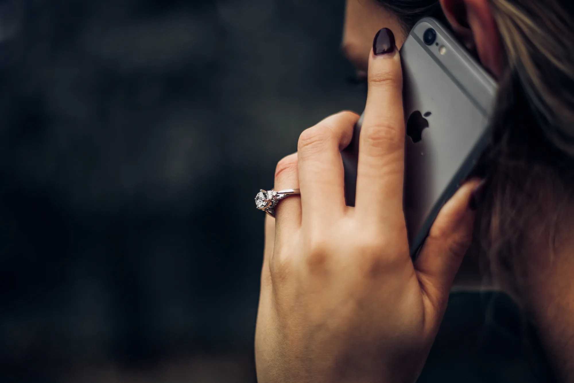 Person with a diamond ring talks on a silver iPhone against a blurred dark background, emphasizing elegance and focus.
