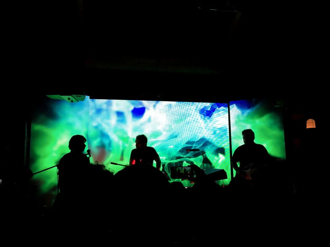 Silhouetted band performing on stage with vibrant green and blue digital backdrop, audience member capturing the moment with a phone.