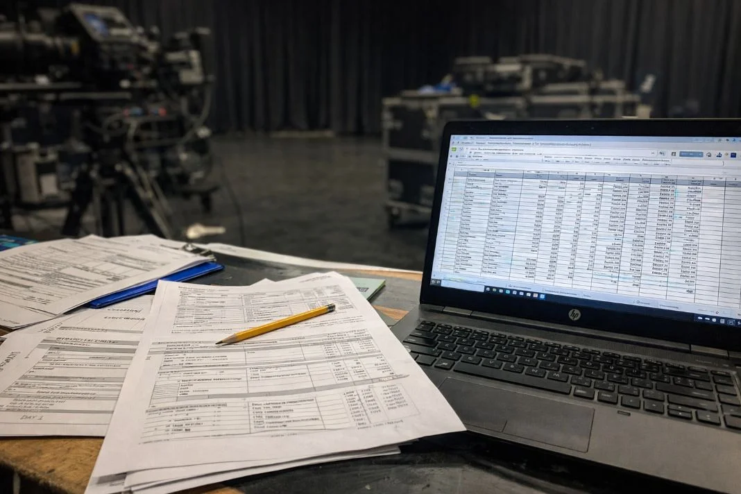 Laptop displaying a spreadsheet and paperwork with a pencil on a wooden table. Background shows film equipment in a studio setting.