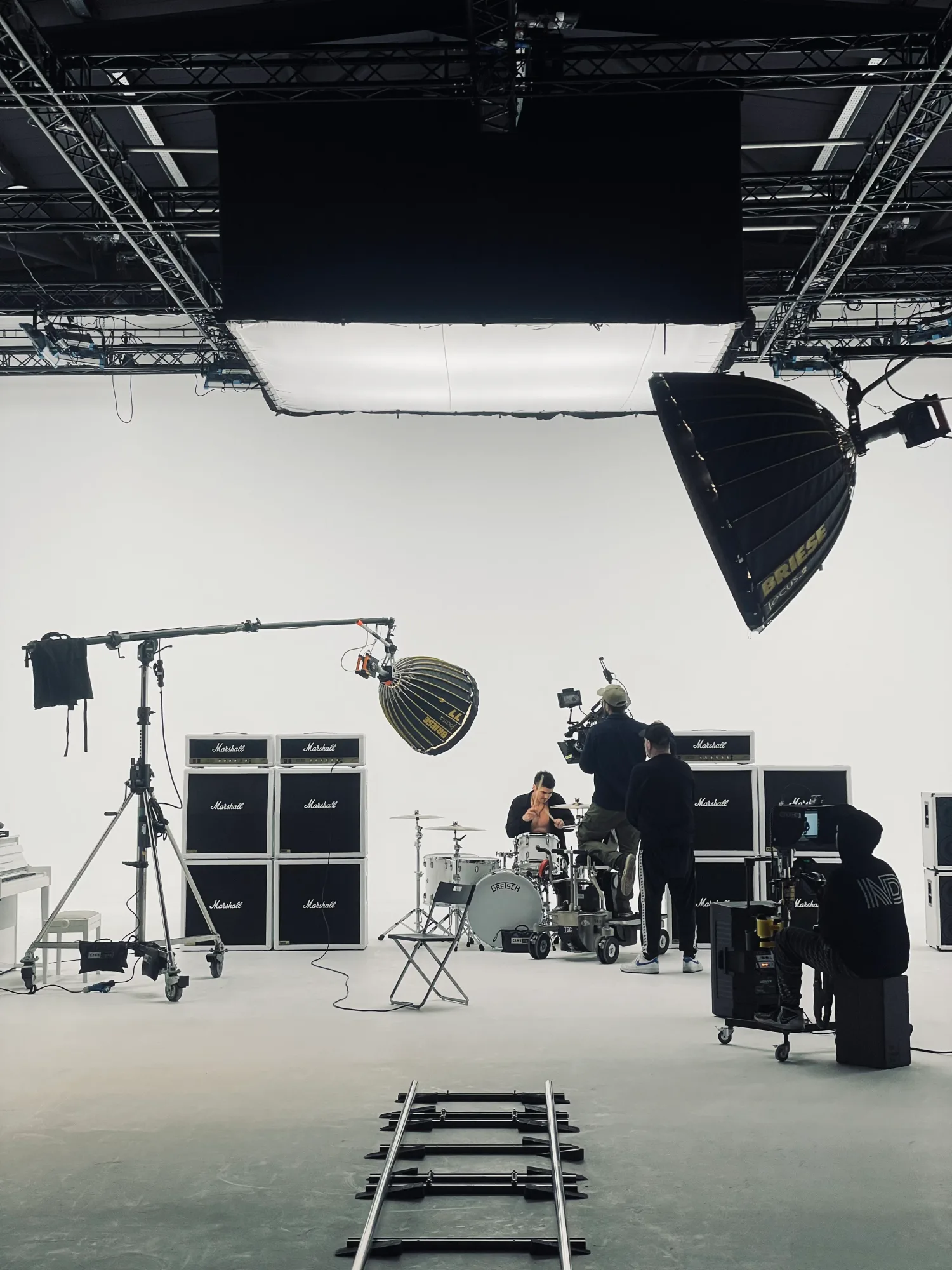 A drummer sits in a bright studio surrounded by Marshall amps. A camera crew films, with lights and tracks visible. Mood is focused.