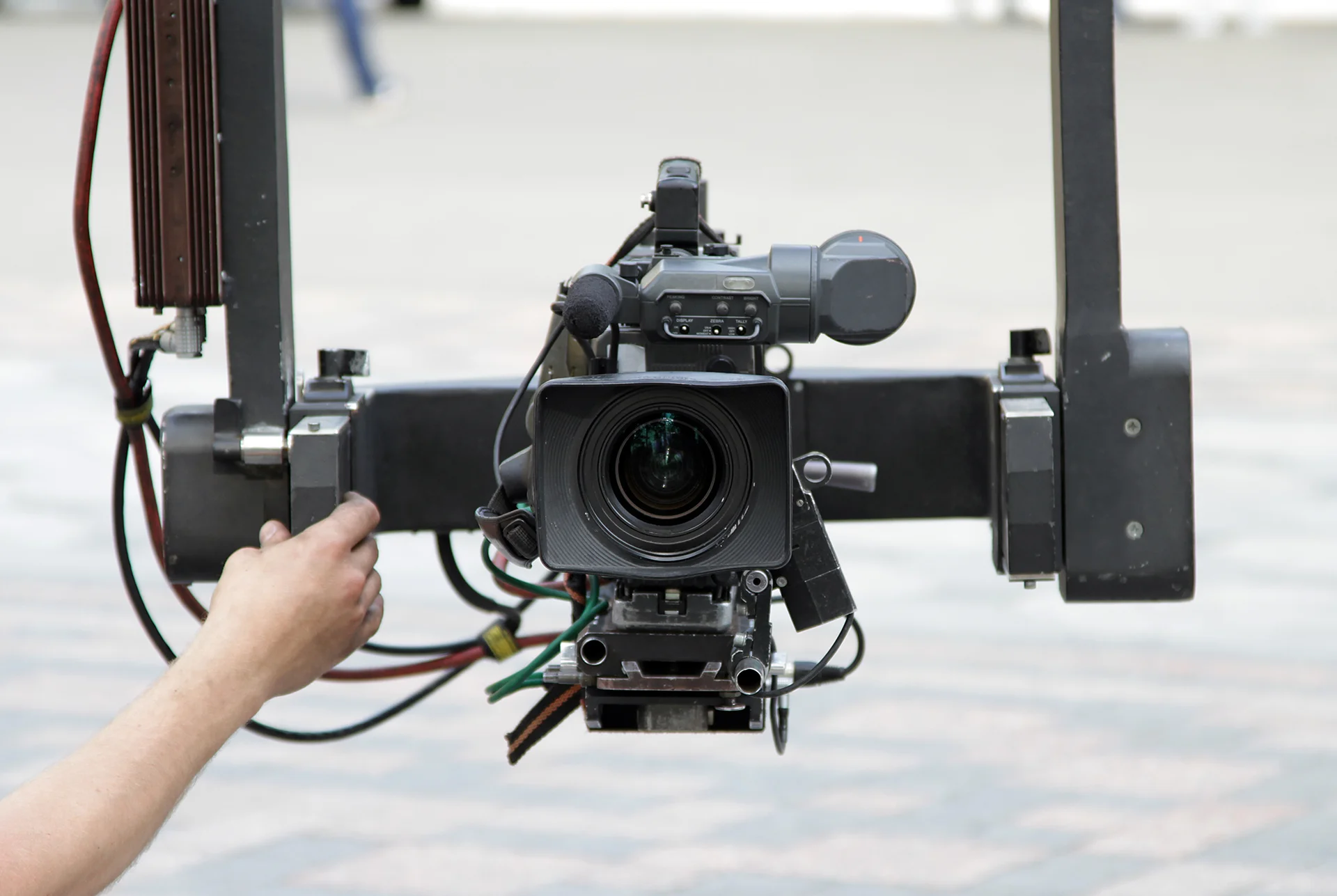 Close-up of a professional video camera on a stabilizer outdoors. A hand adjusts the equipment. Background is a blurred paved area.