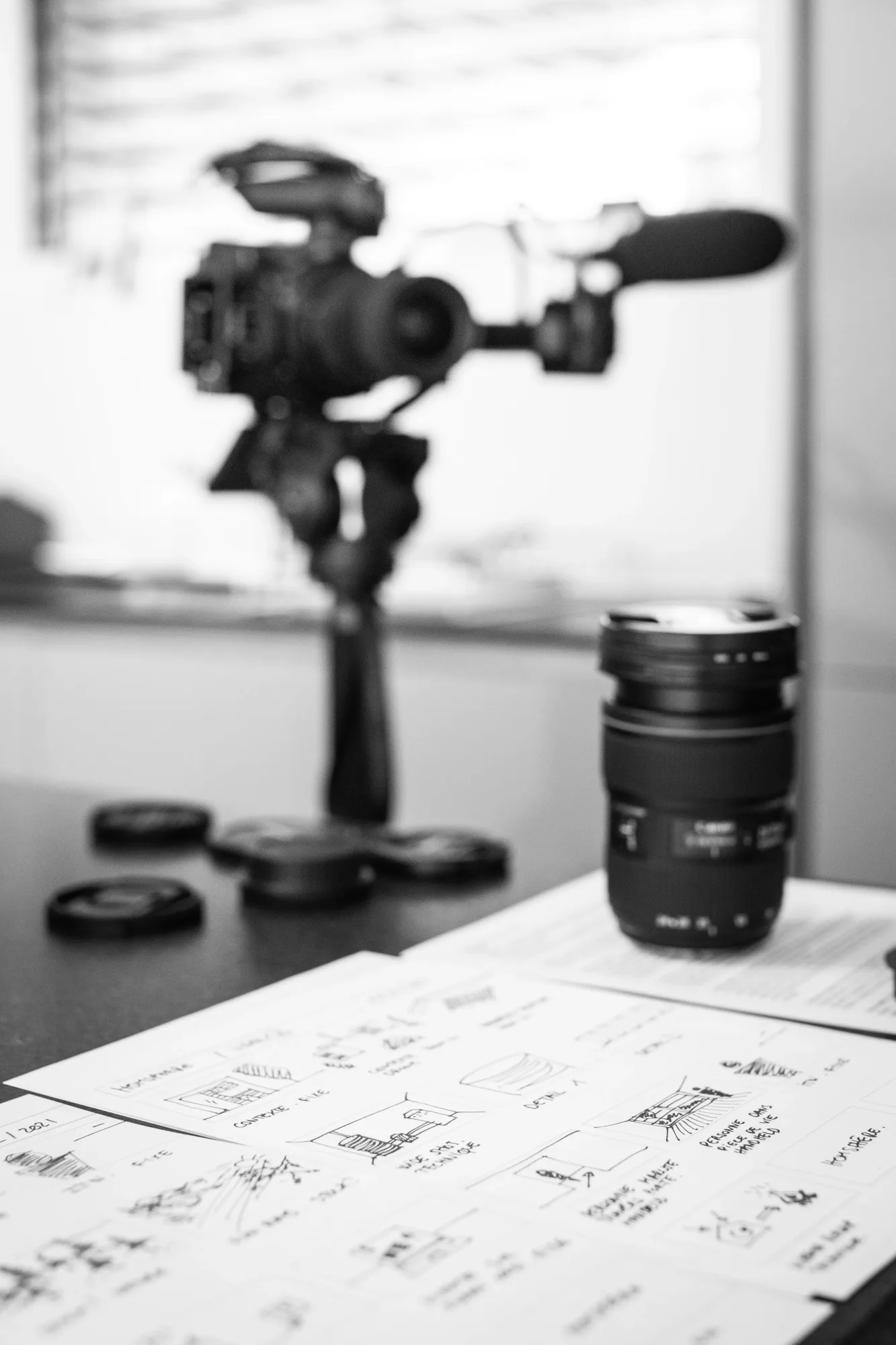 Camera and lens on a table, surrounded by scattered lens caps. Storyboards with sketches and text in the foreground. Monochrome setting.