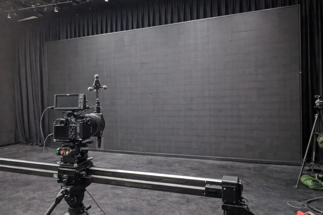 Camera setup on slider in a studio with black curtains and large LED screen. Moody, dimly lit atmosphere, no visible text.