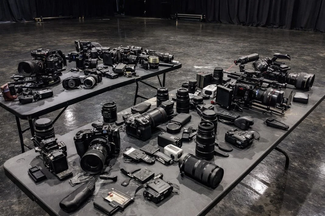 Tables filled with various cameras and lenses on a dark glossy floor set. The background features a curtain, creating a moody atmosphere.