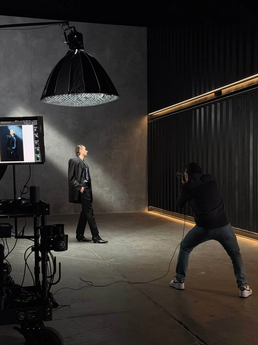 Photographer captures a model in a sleek suit, hands in pockets, under studio lighting. Gray walls and screen display his portrait.