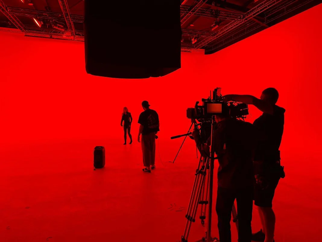 Camera crew films two people on a red-lit set. Silhouettes and intense lighting create a dramatic atmosphere.