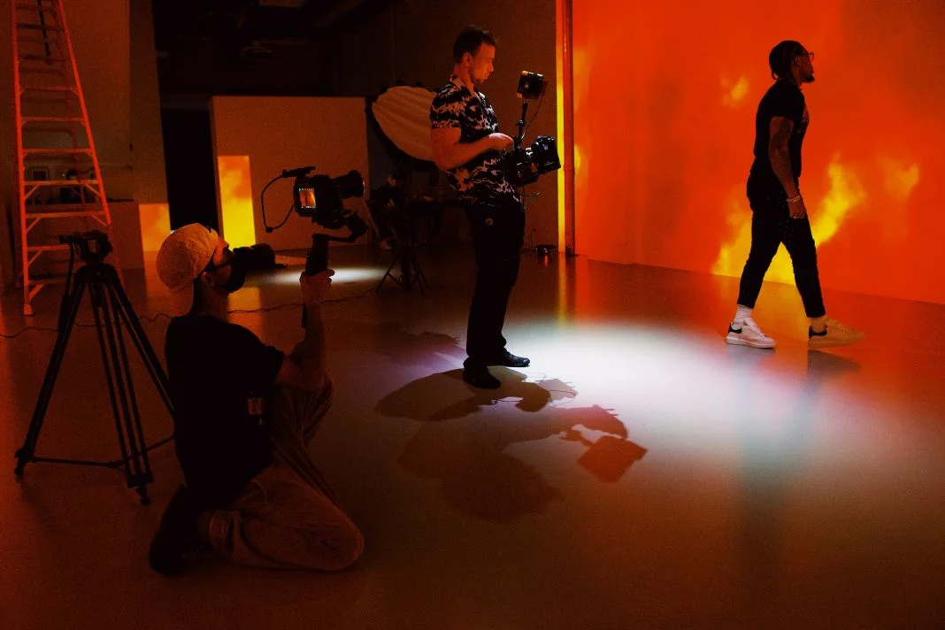 Three people in a studio with orange lighting. One kneels with a camera, another films, and a third walks. A ladder and tripod in view.