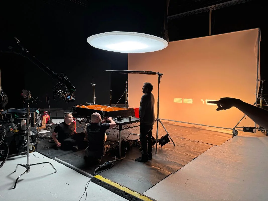 Film set with three people adjusting equipment under a large light. An orange backdrop and a pointing hand suggest active preparation.