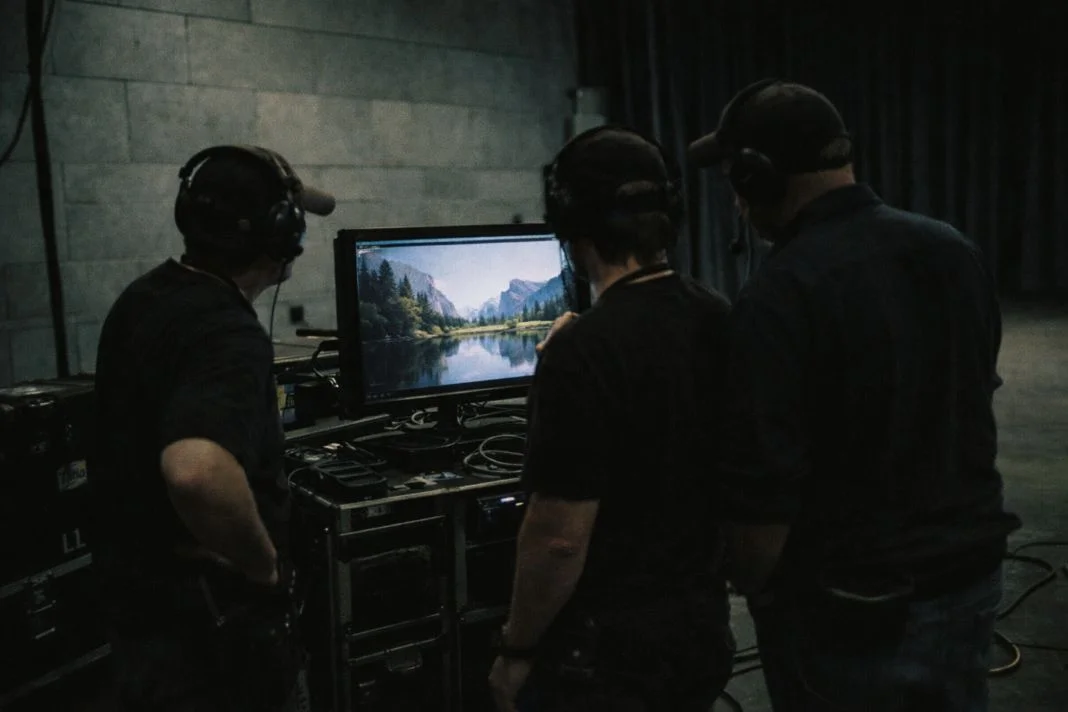 Three people with headsets view a monitor displaying a scenic landscape. They stand in a dim room with industrial walls and equipment.