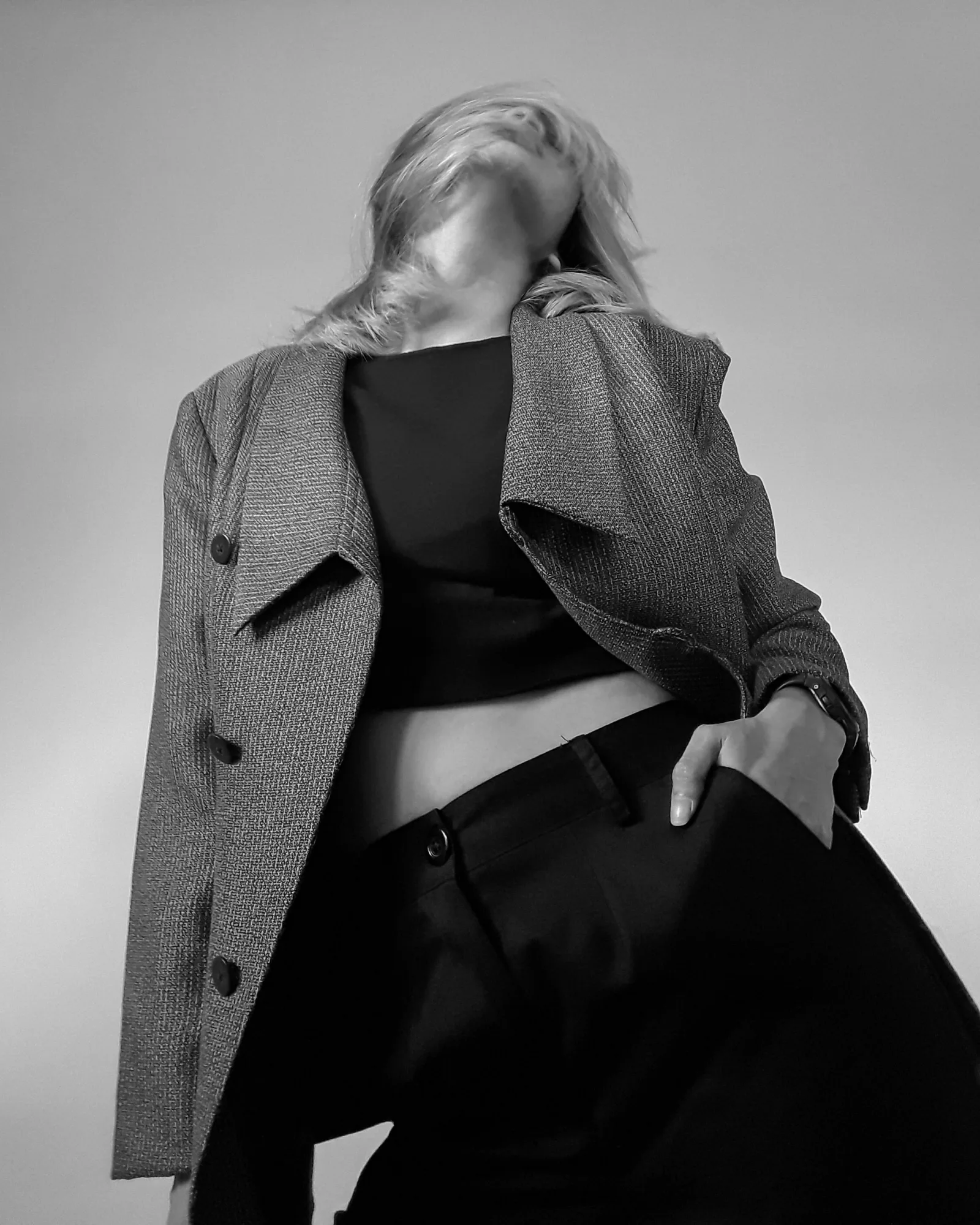 Low-angle shot of a person in a gray coat and black outfit, head tilted back. Monochrome setting, evoking a confident mood.