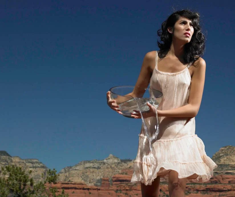 A woman in a white dress holds a glass bowl with water spilling over. She stands against a backdrop of red rock formations under a clear blue sky.