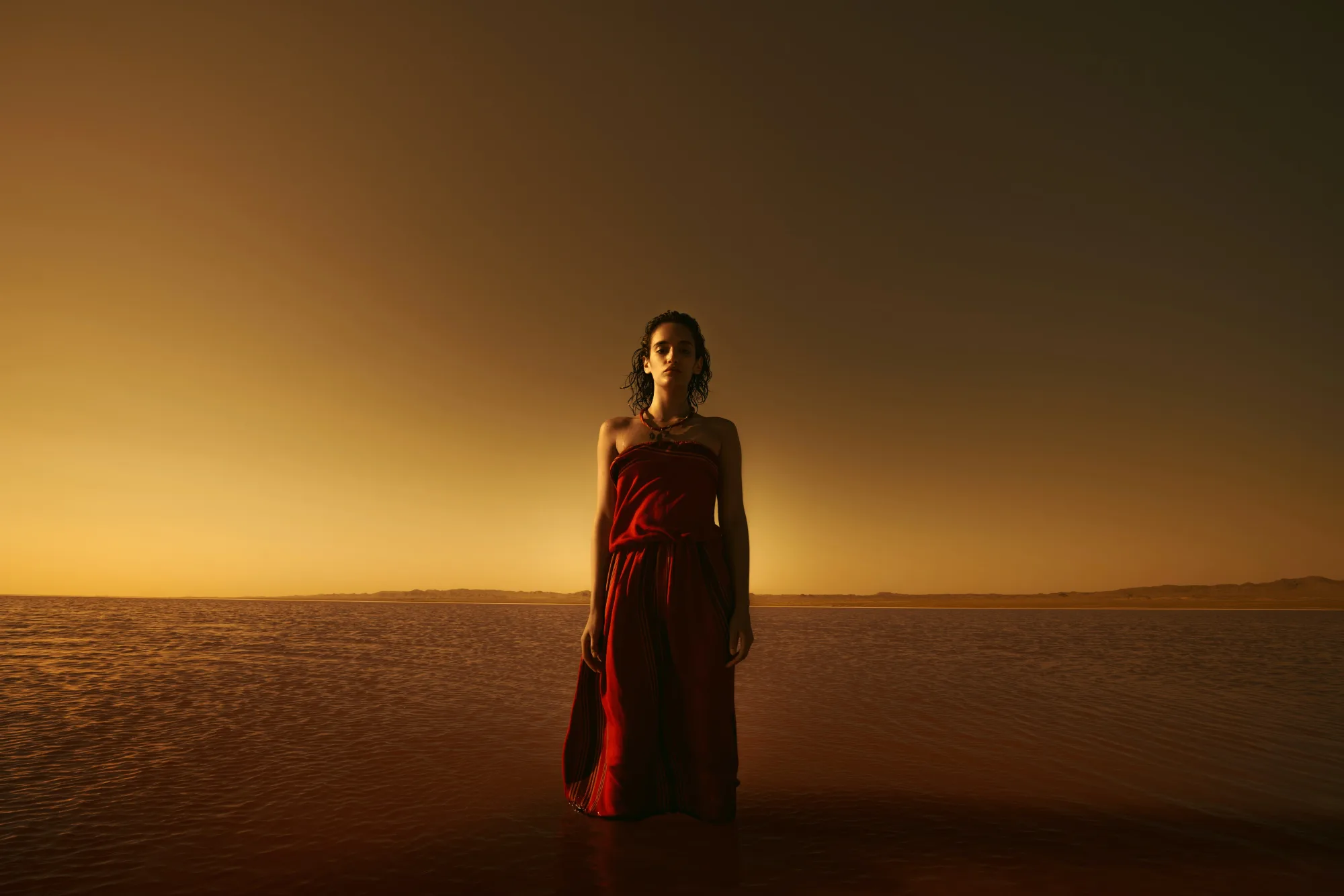 Woman in a red dress stands in calm water at sunset, with a serene expression. The sky and water glow in warm, golden hues.