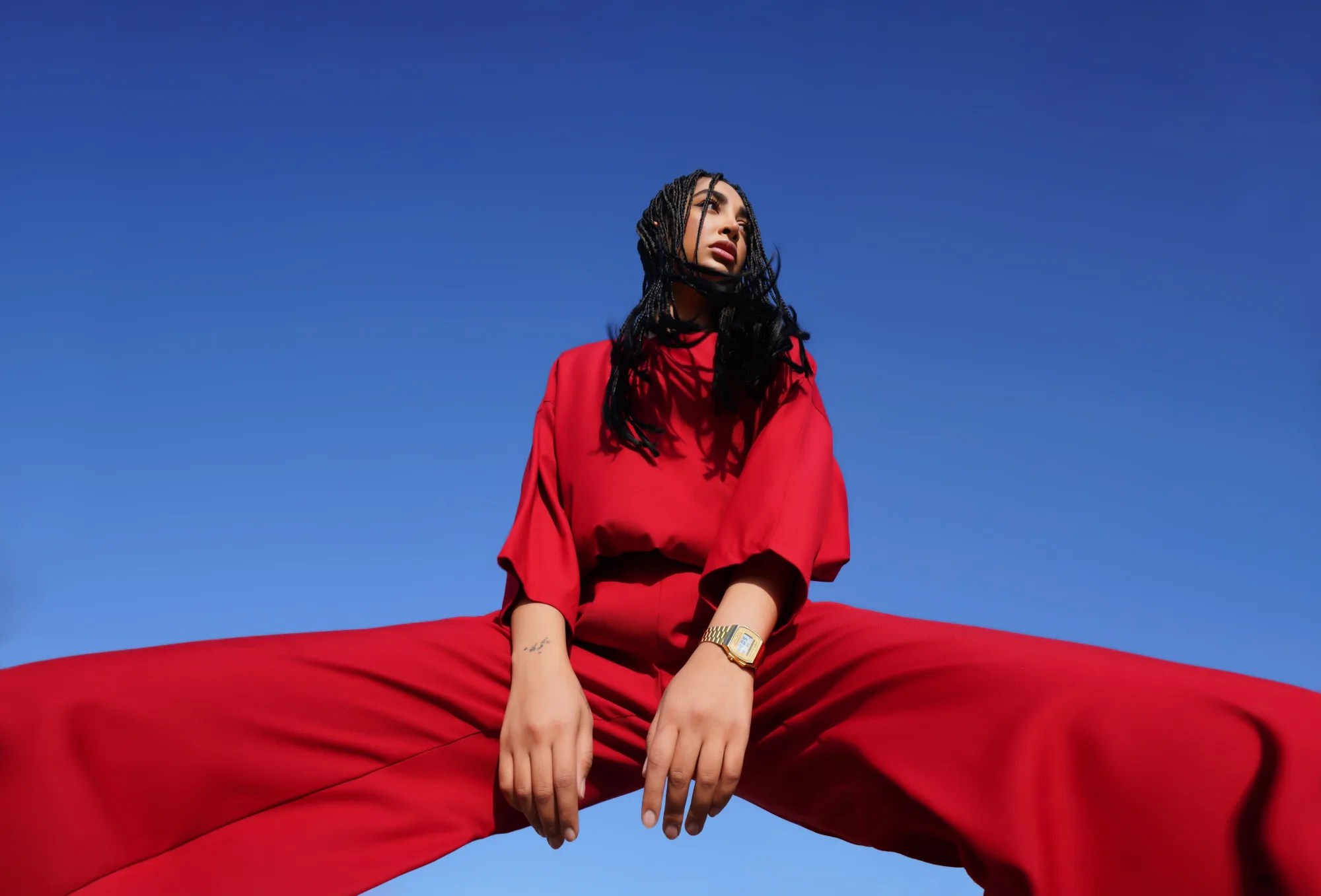 Person in red outfit poses against clear blue sky, legs wide apart, with braids and gold watch. Confident and bold mood.