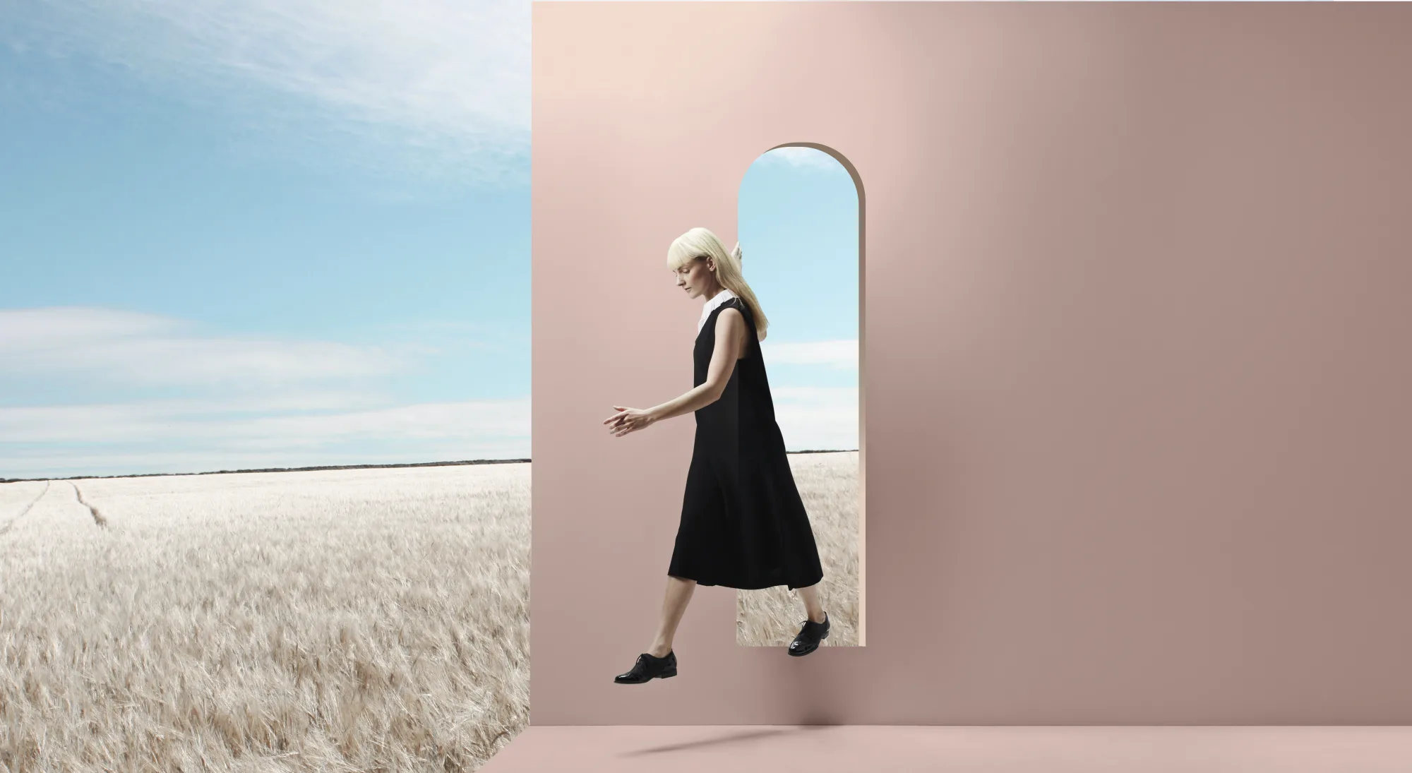 Woman in black dress appears to walk through a pink archway into a wheat field under a blue sky. Surreal and serene atmosphere.