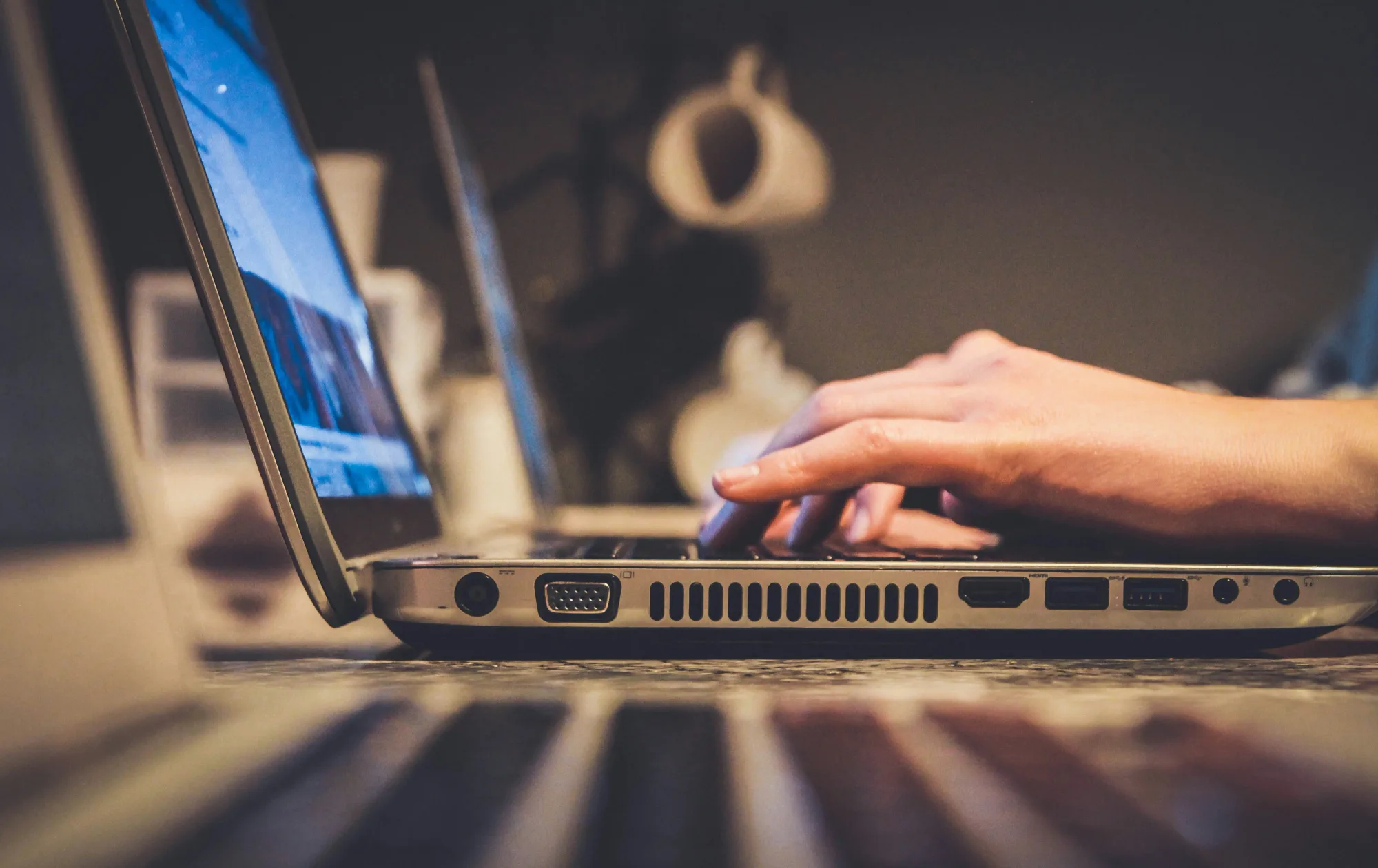 Hands typing on a laptop keyboard, focus on fingers. Blurred background features another laptop and white objects. Warm, cozy ambiance.