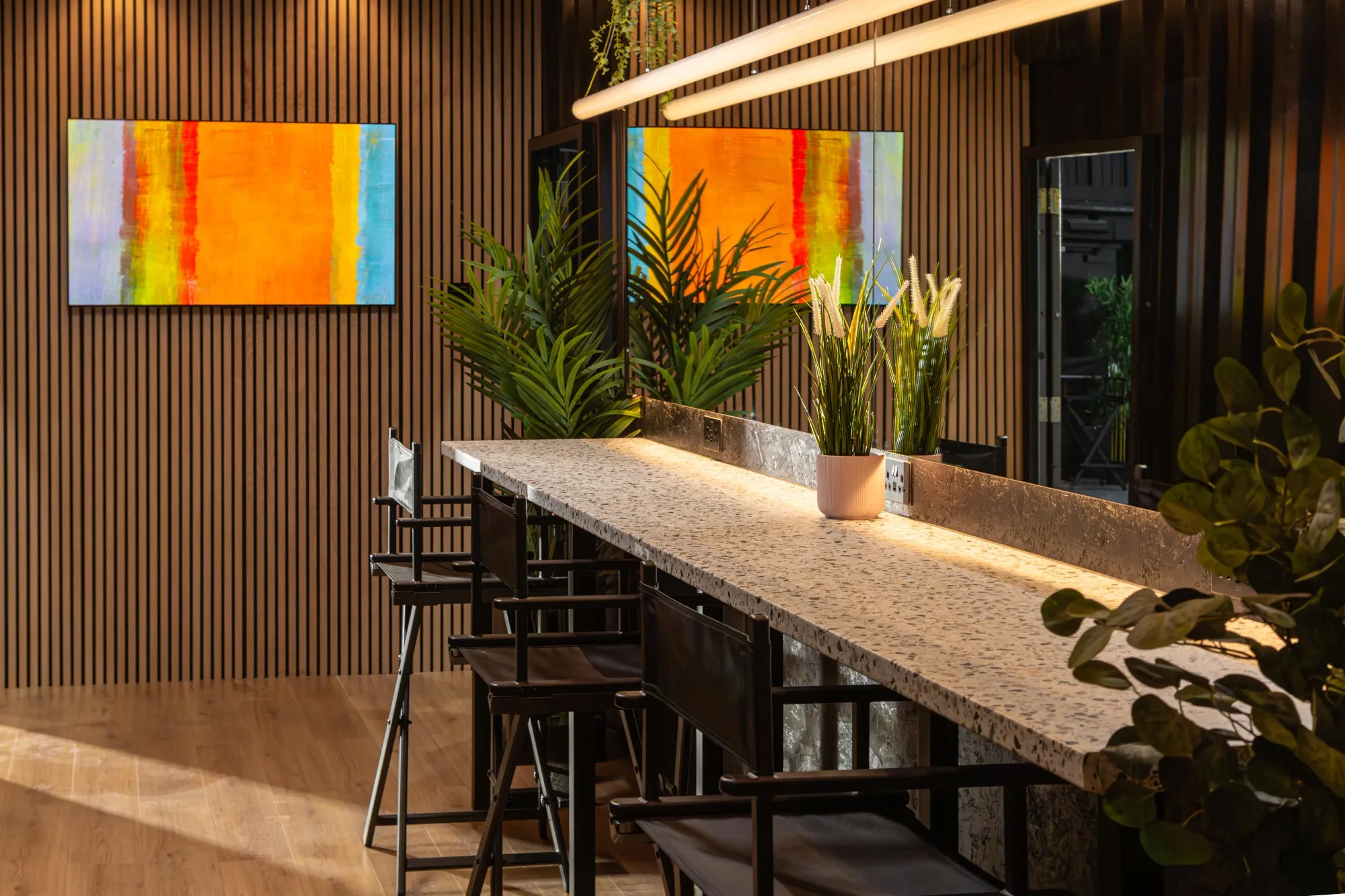 Modern seating area with terrazzo table, chairs, vibrant abstract art, and plants. Vertical wood paneling and warm lighting create a cozy ambiance.
