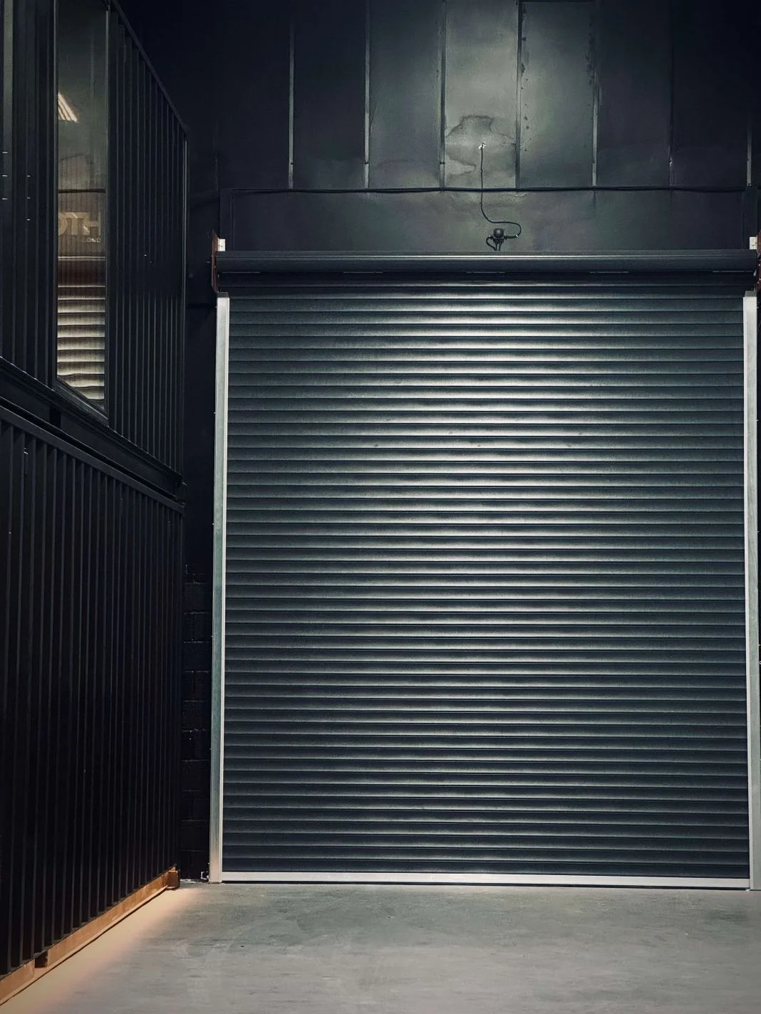 Black metal roll-up door in an industrial setting. Dark walls and floor create a minimalist atmosphere. No visible text or people.